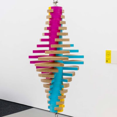 A wooden sculpture suspended from the ceiling, with a central cylinder and shell-like oval bulge in the middle. It is designed to rotate, and is painted maroon, cyan and yellow, which blur together to white when the sculpture is rotating fast enough.
