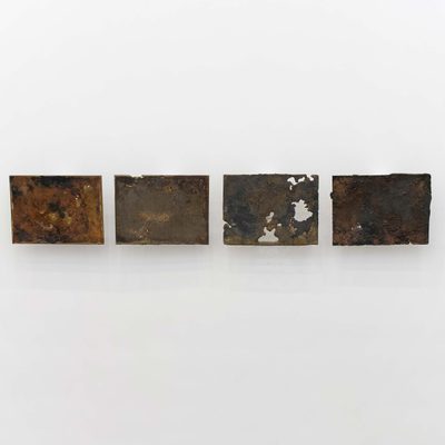 Four rectangular pieces of cast bell bronze, hung in a row on a white gallery wall. They have rough, pockmarked surfaces and occasional holes, giving them the appearance of topographical maps or aerial photography.