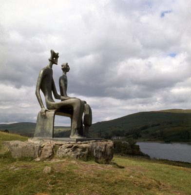 A sculpture of a seated King and Queen sited on a hilltop overlooking a lake. The sky is cloudy with shadows crossing the lake.