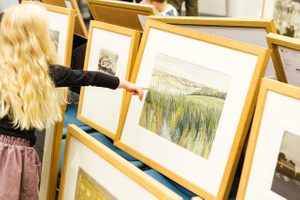 A young girl with long blond hair points to a framed painting in a crowded selection of frames.