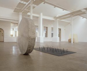 A large, white, disc-shaped sculpture stands next to a flat, floor-based sculpture with many protrusions rising out of it like twigs.