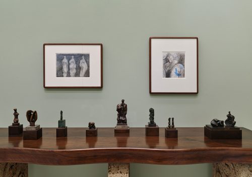 Eight small bronze sculptures by artist Henry Moore, displayed on a wooden table. Two framed drawings, also by Moore, are on the wall behind.
