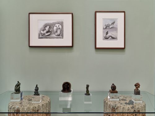 Six small bronze sculptures by artist Henry Moore, displayed on a glass table. Two framed drawings, also by Moore, are on the wall behind.