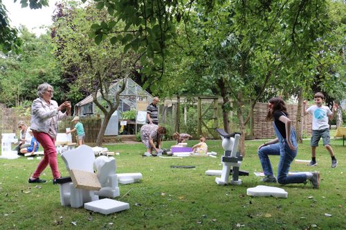A group of adults and children building sculptural shapes using soft foam blocks and a range of recycled materials. They are all working in a small orchard next to a greenhouse.