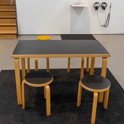 A small rectangular table with four round stools on a piece of carpet in the middle of a gallery space.