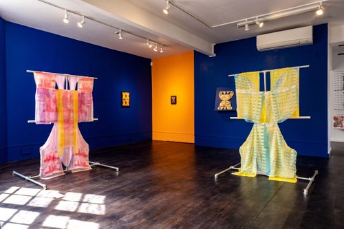 An art gallery with blue and yellow walls, displaying textile sculptures displayed on metal frames.