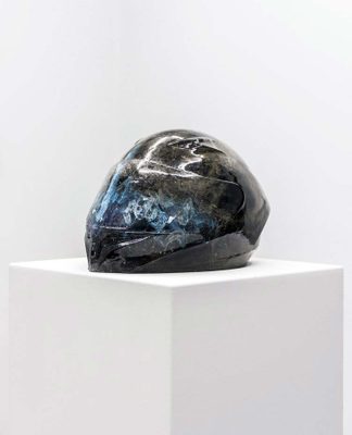 A black, glass sculpture of a motorcycle helmet, displayed on a white plinth.