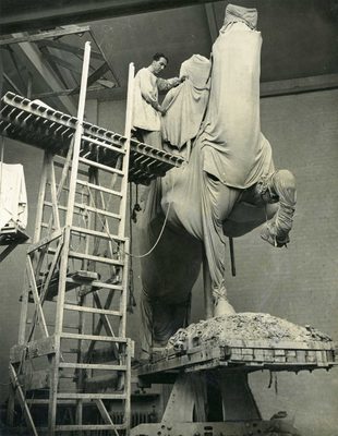 Sepia photo of a man stood high up on a wooden platform so that he can work on the upper part of a monumental sculpture. The sculpture is covered by sheets, but it is clear that it depicts a horse and rider.