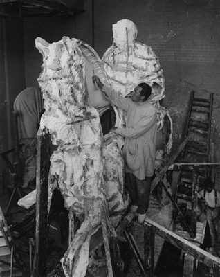 Black and white photo of a man stood high up on a wooden beam so that he can work on the upper part of a monumental sculpture. The sculpture is still early in development, but it is clear that it depicts a horse and rider.