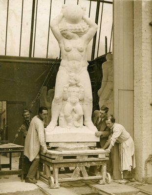Sepia photo of four men carefully moving a large stone sculpture of a woman and child by means of a wooden platform with wheels.
