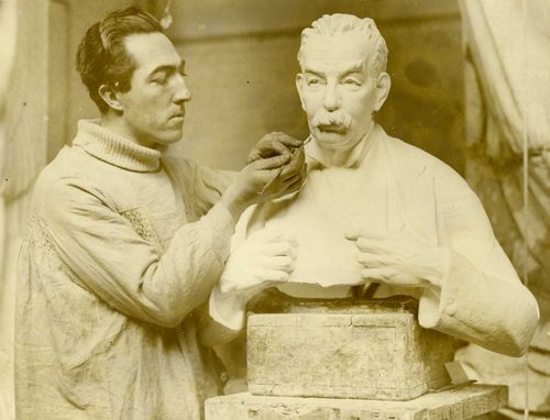 Sepia photo of a man posing at work by a finished plaster portrait bust of a man with a prominent moustache.
