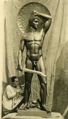 Sepia photo of a man kneeling down to put the finishing touches to a sculpture of a muscular male nude, standing at around nine or ten feet tall. The figure carries a sword in its right hand, and with the left it holds a round shield with decorative inlay above and behind its head.