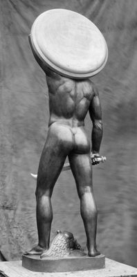 Black and white photo of a muscular nude man, seen from behind. He carries a sword in this right hand, and holds a raised shield behind his head with his left.