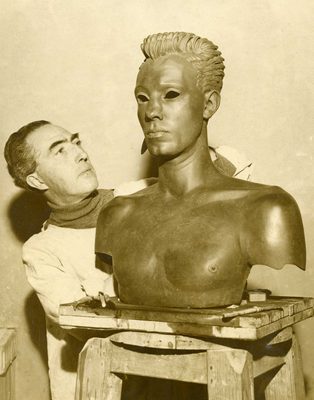 Sepia photo of a man putting the finishing touches to a male portrait bust, slightly larger than life size. The face of the sculpture has no eyes, and through the eye-holes it is evident that the sculpture is hollow.