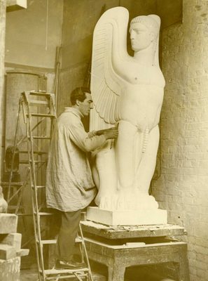 Sepia photo of a man putting the finishing touches to a large plaster sculpture of a sphinx in the Greek tradition, with the head of a man, body of a lion and wings of an eagle.