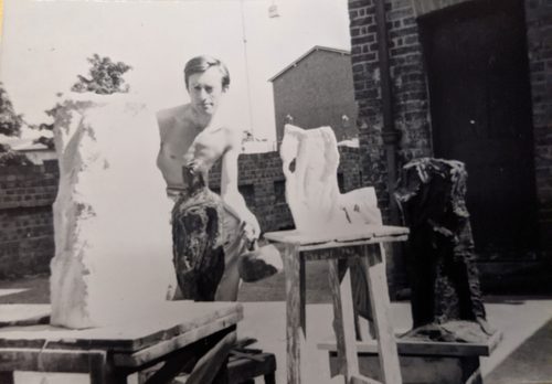 Black and white photo Anthony Hatwell, who is working topless outdoors carving a series of small sculptures.