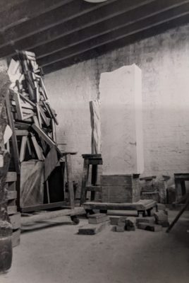 Black and white photo of the interior of a warehouse-like space. On one wall pieces of wood look to be joined together haphazardly, reaching up to the ceiling. In the centre of the photo there is a block of stone, ready to be carved.