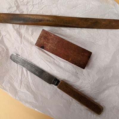 Old, well used tools for sculpting, comprising a wooden-handled knife with a wide, flat blade; a rectangular wooden block; and a long, rounded modelling tool with 'Hatwell' scratched into it.