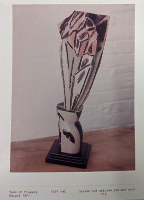 Abstract sculpture by Anthony Hatwell, carved from wood to give the impression of tall flowers, clustered closely together, stood in a small vase. It is painted white with black lines, with the petals of the flowers a light pink.