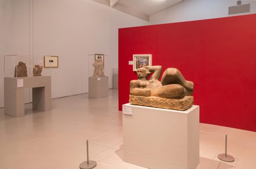 Sculpture of woman laid on her side, with one arm behind her head, carved from stone by Henry Moore. The sculpture is on a plinth, and behind is a free-standing red wall, with other sculptures around the room.
