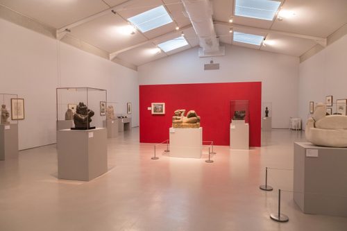 A large gallery space with a free-standing red wall in the centre of the room. Displayed on plinths around the room and many figurative sculptures by Henry Moore, carved from wood and stone.