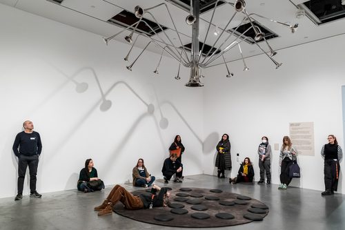 A sculpture installation in a gallery. A group of people are gathered around a circular carpet on the floor, which has several large circular objects like game counters on it. From the ceiling a large metallic abstract shape, made from trumpet like funnels, extends down towards the carpet.