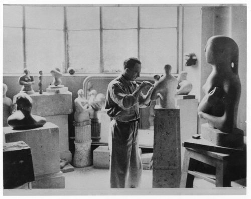 A black and white archive photo showing a sculptor, Henry Moore, aged in his 30s, working in his studio. He is working on a small stone sculpture of a Mother and Child
