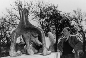 A black and white photo of a man, in a sweater vest and slacks, he is cleaning a reclining figure sculpture on a plinth.