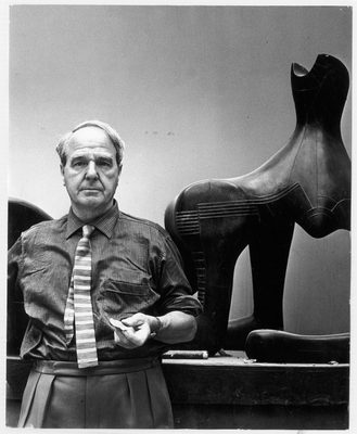 A black and white photo showing a middle aged man, in shirt and tie, standing in front of an abstract sculpture which depicts a figure reclining