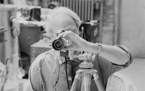 A black and white photo of Henry Moore, who is sitting behind his own Leica camera on a tripod as if taking a shot.