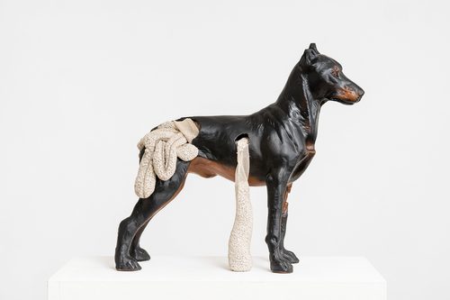 A ceramic sculpture of a brown dog standing upright and staring to the right. Soft cream gauze bandage spills from its stomach and back.