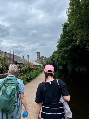 Photo of two adults walking along a canal path. Mature green trees overhang the canal to the right, and large stone building, probably a former mill, can be seen up ahead.