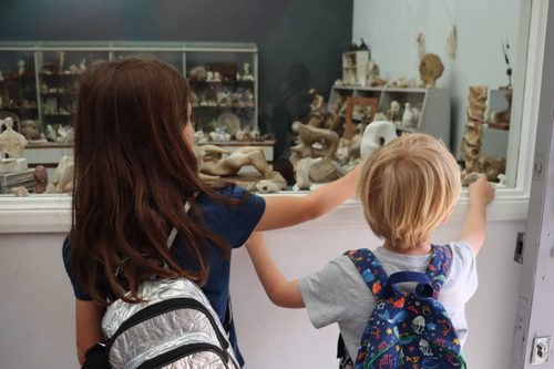 A dark haired young girl and a blonde boy stand looking through a window into an artist's studio which is filled with small sculpture models