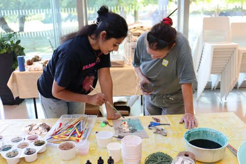 Two women, one older one young, are working at a table using a paint brush to apply colour and metallic coating to a piece of art.