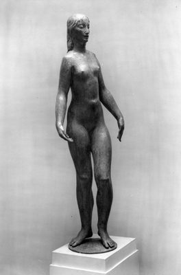 Black and white photo of a bronze sculpture of a slim female nude. The figure is standing in a relaxed pose, with the left leg slightly further forward and the heel raised, as if she is about to take a step forward. The sculpture is shown indoors against a white wall, mounted on a white plinth.