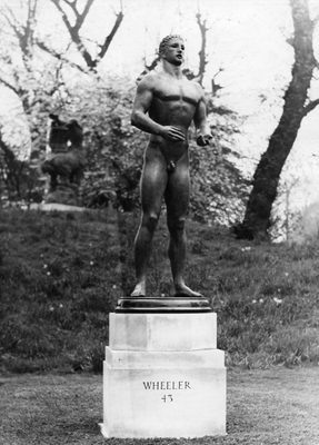 Black and white photo of a bronze sculpture of an idealised male nude. The figure is standing, with elbows bent and hands raised to just above the hips. The sculpture is shown outdoors, mounted on a white stone plinth in which the words 'WHEELER 43' are carved.