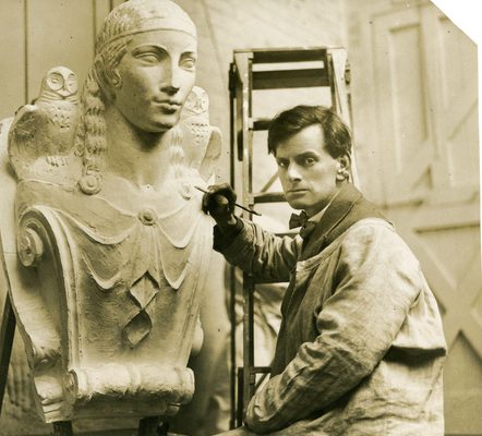 Sepia photo of a white man with brown hair working on a plaster sculpture. He is wearing a suit and bow tie, with overalls over the top, and there is a ladder behind him. He holds a modelling tool in his right hand, which is raised up close to the sculpture. He is twisted away from his work to look directly at the camera. The sculpture he is working on is a larger-than-life size portrait bust of a woman. There is an owl perched on each of her shoulders. The sculpture looks architectural in nature, like it will adorn the front of a building.