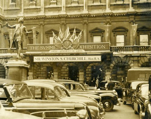 Sepia photo of the front of the Royal Academy, a large brick built building, taken in the 1950s. Many cars are parked outside. A sign on the front of the building reads 'The Summer Exhibition | Royal Academy of Arts | Sir Winston S. Churchill. Hon. R.A.'.