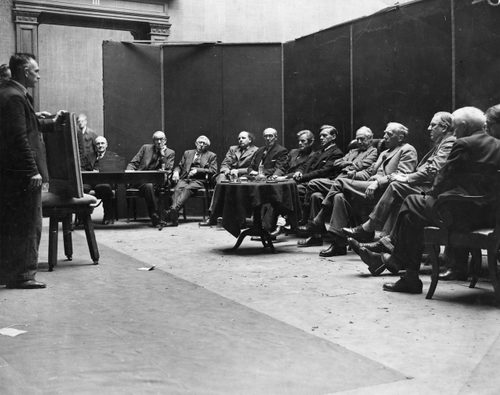 Black and white photo of a room full of old white men. On the left, two men stand holding up a painting that is resting on a stool; it is facing away from the camera, so we can't see what is painted on it. Thirteen other men are sat in a semi-circle around, judging the painting.