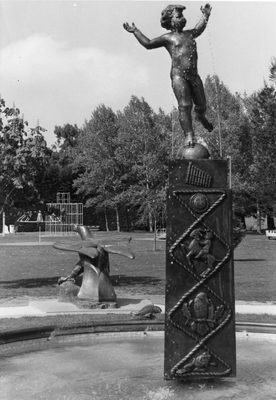 Black and white photo of a fountain sculpture. It comprises a naked child with arms raised, balanced on its left leg on top of a ball. The ball is mounted on a tall, square column, which is decorated with various relief sculptures (from top to bottom): a pan flute, a face, a child riding a goat, a bird, a frog. Behind the fountain, there is a large sculpture of a duck facing away, with a climbing frame behind that, and trees in the distance.