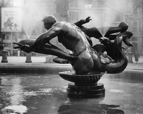 Black and white photo of a fountain sculpture. It comprises a merman leaning forwards, holding a dolphin in his outstretched left hand, while another fish, small dolphin, and young mer-person swim around him.