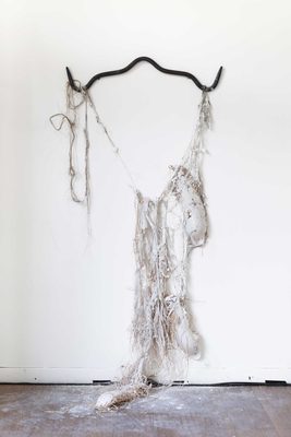 Abstract sculpture made from white kaolin clay coating coarse raffia fibres, hanging from an iron gambrel attached to a wall.