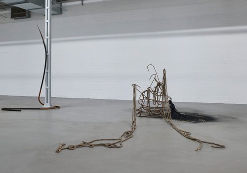 Abstract sculpture in a white gallery space. It is made from rusted metal harpoons and rope, arranged in a vaguely conical shape.