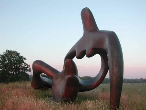 A huge abstract sculpture of a reclining figure, sited on a hill. It is sunrise and pink light is washing over the sculpture.