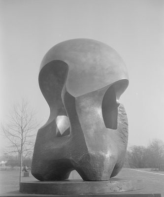 An abstract bronze sculpture, photographed outdoors. The top is smooth, curved and highly polished, while the lower parts and rougher, with scratches and gouges still evident where tools have been used.