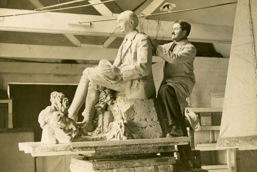 Sepia photo of a man with dark hair and a moustache working on a life-sized sculpture of a seated man.