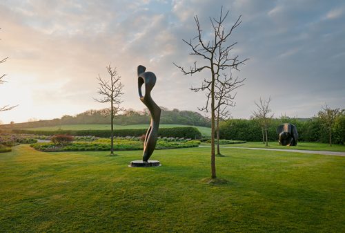 The setting sun illuminates a towering bronze sculpture of an abstract standing figure by Henry Moore, displayed on a manicured lawn in an English sculpture garden.