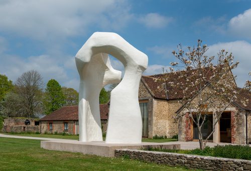 A large, arch-shaped sculpture by Henry Moore, displayed on the grounds outside a stone-walled building that has been converted into a gallery. The arch's shape and chalk-white colour gives the impression of bones.