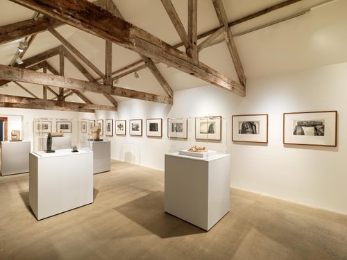 A white-walled gallery space with exposed wooden beams, showcasing work by Henry Moore. Framed drawings are hung on the wall, and there are small sculptures displayed underneath perspex containers on five plinths.