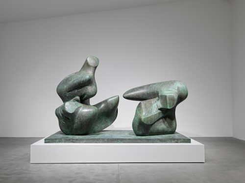 Large bronze sculpture by Henry Moore of an abstract reclining figure, split into two pieces at the torso. It is displayed on a plinth in a white-walled gallery space.